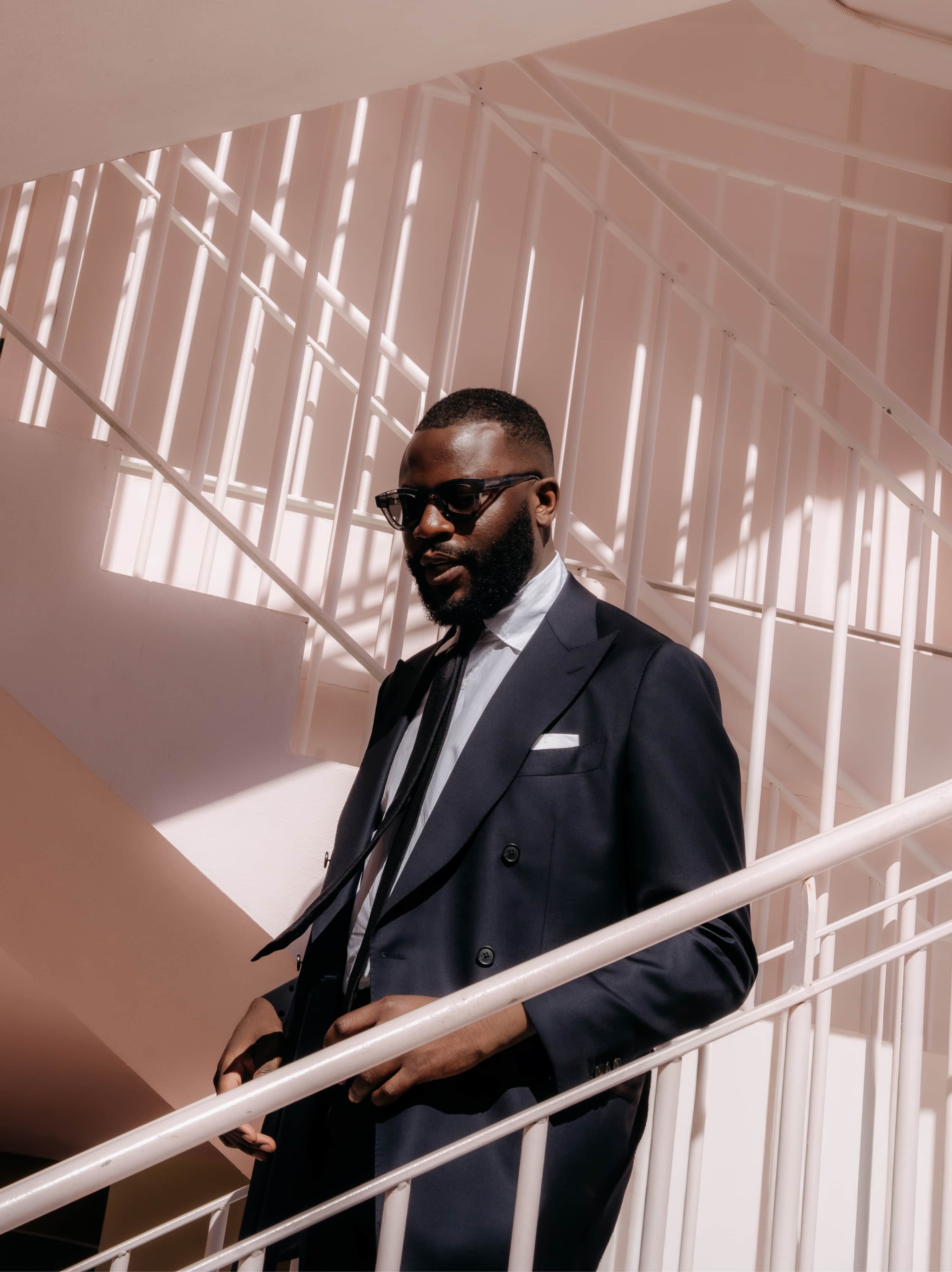 A model wearing dark sunglasses and a summer navy suit from Suitsupply is going down pale pink stairs