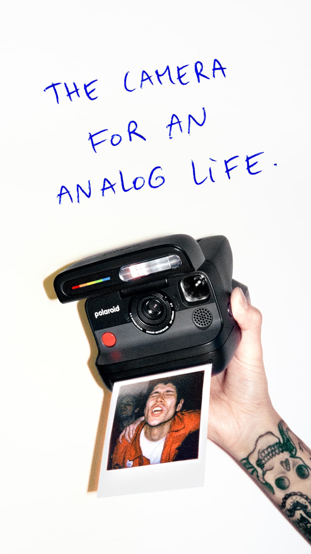 A tattooed hand is holding a black Polaroid Flip camera with an instant film showing a fun portrait. Above is handwritten ' The camera for an analog life'