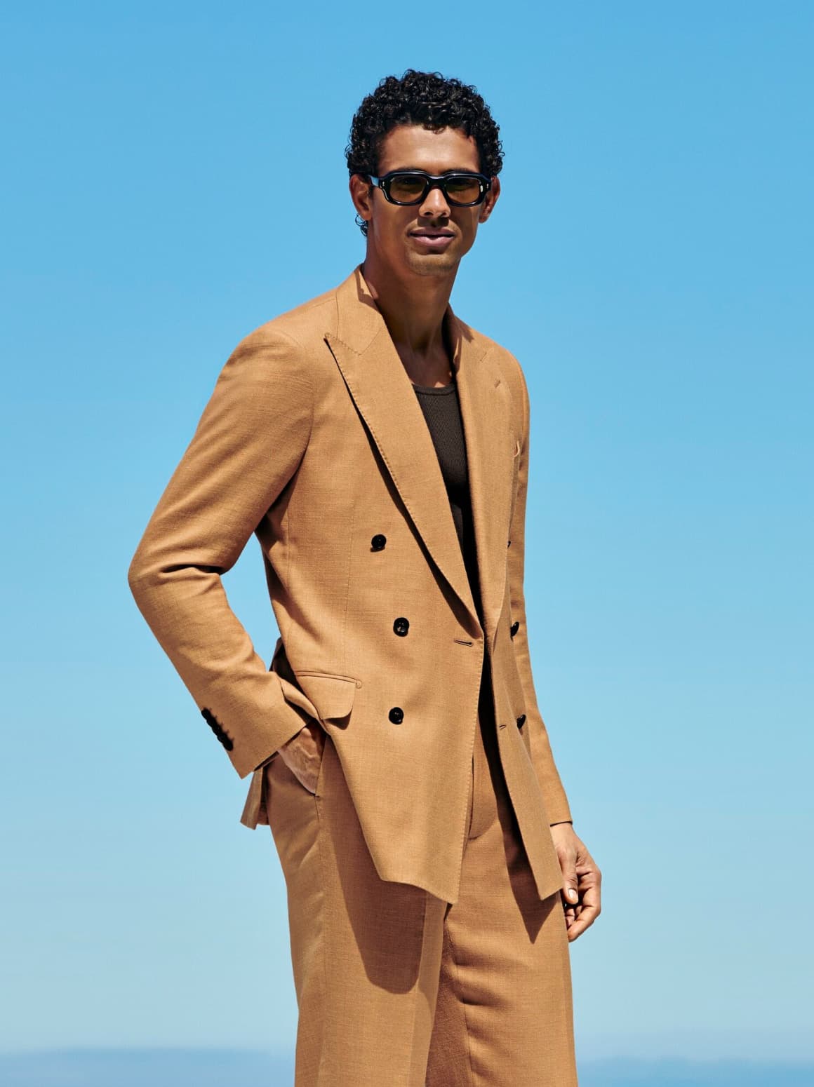 A person with black sunglasses and one hand in their pocket is wearing a camel summer Suitsupply suit over a blue sky horizon.
