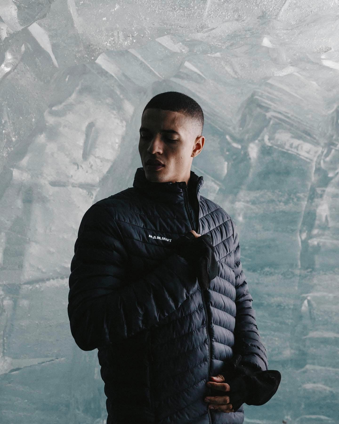 A person wearing a dark blue Mammut jacket stands in front of a textured ice wall, looking thoughtfully to the side.