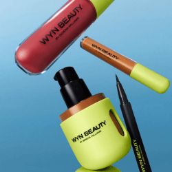 Assorted WYN Beauty makeup products, including lip gloss, concealer, foundation pump bottle, and liquid eyeliner, against a blue background.