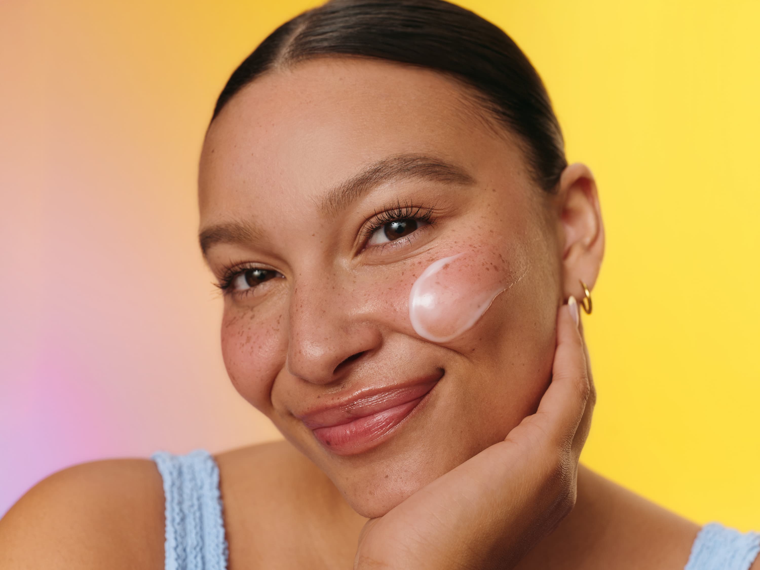 A close-up of a person's face smiling with some Jergens Hydra Gel on their left cheek over a gradient pink to yellow background.
