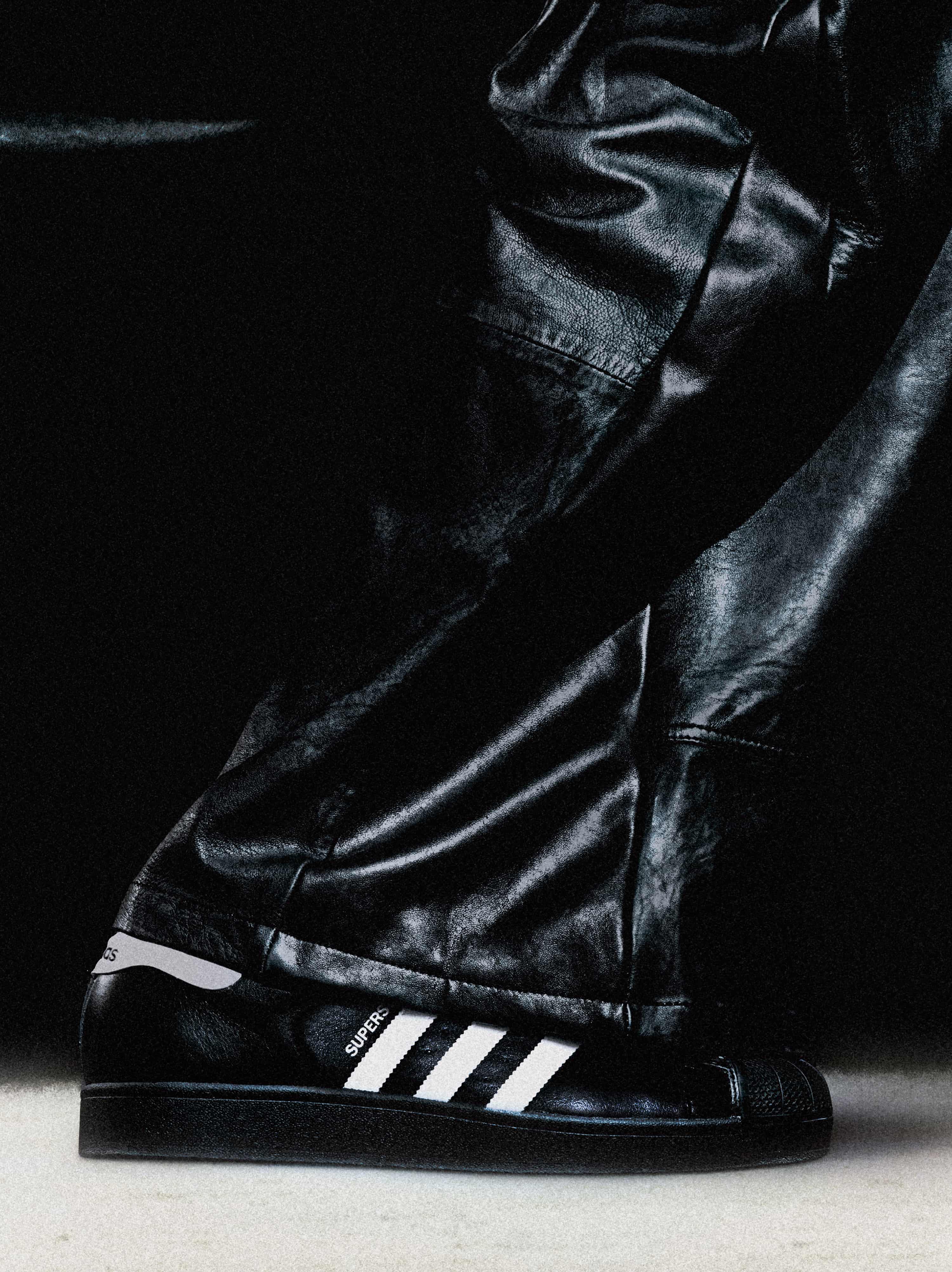 A closeup of a leg wearing a leather trousers and the iconic Superstar shoe model from Adidas in black and white.