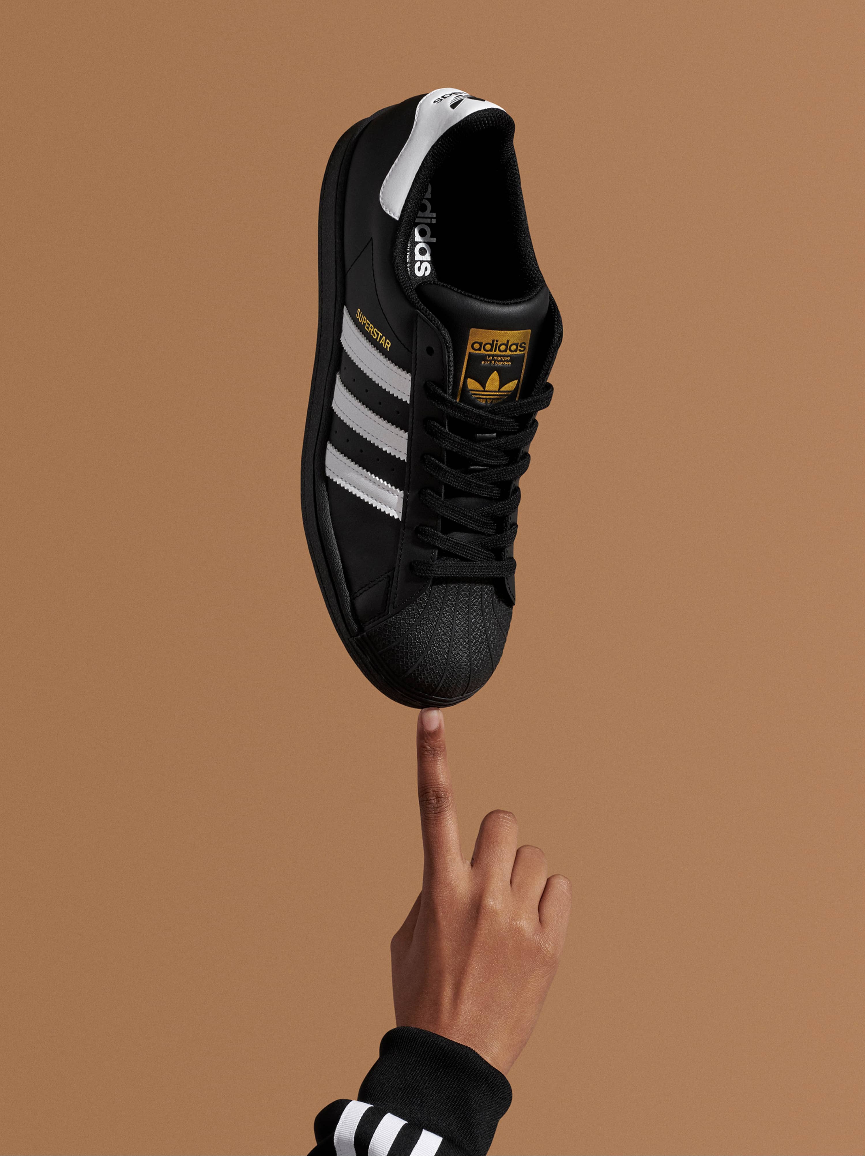 A Superstar Adidas black and white shoe is balancing on a hand over a brown background