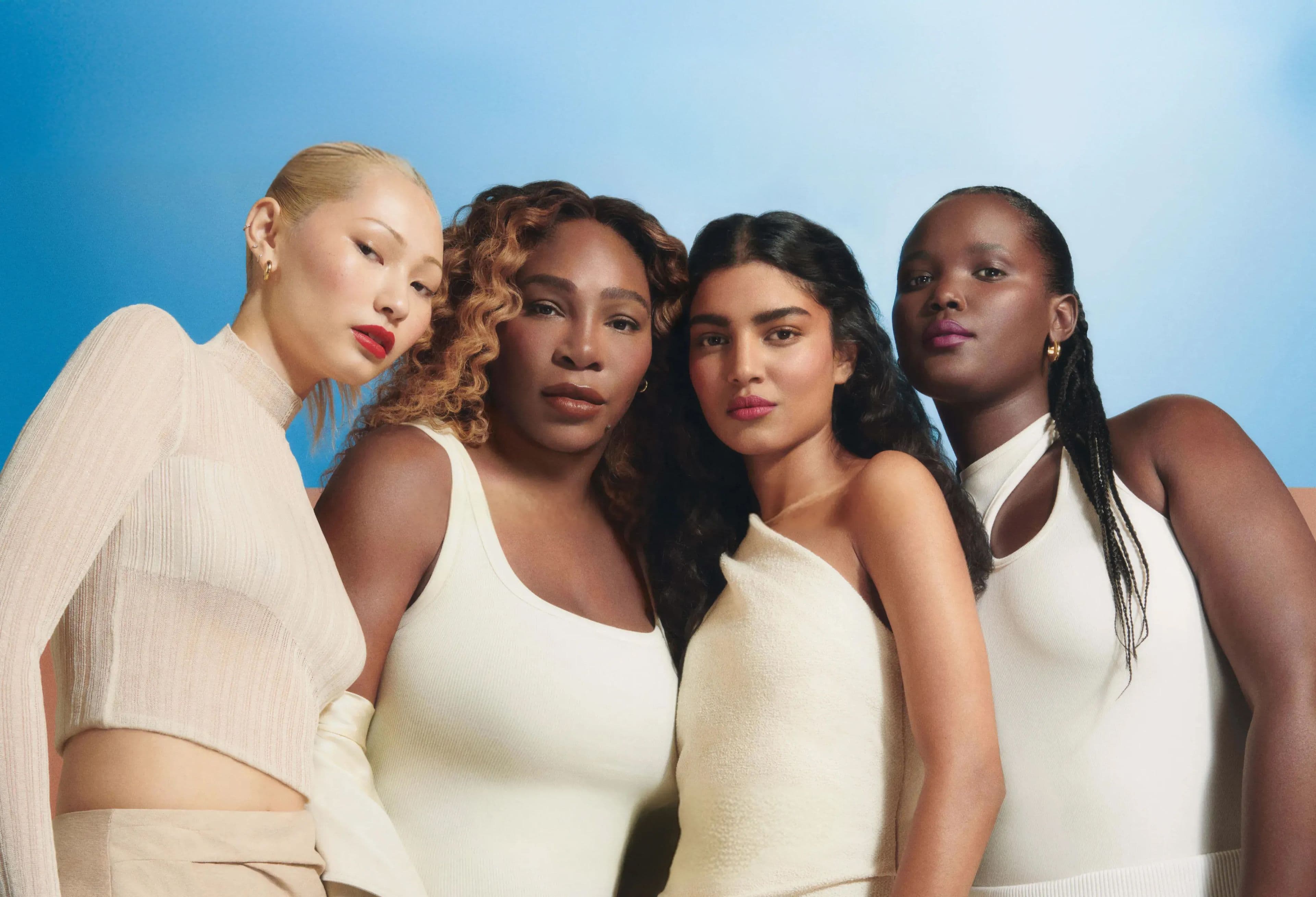 Four people of diverse skin tones stand closely together against a soft blue gradient background, wearing coordinated cream and white sleeveless and knit garments. Each person wears subtle gold jewelry and natural makeup with bold lip colors, creating a modern, inclusive beauty campaign with clean styling and confident expressions.
