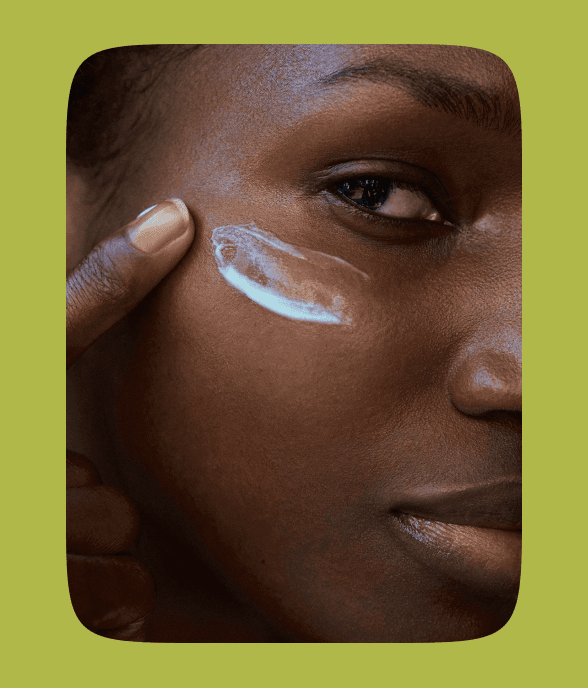 Person applying a streak of cream under their eye on smooth, dark skin, with a focused expression against a green background.