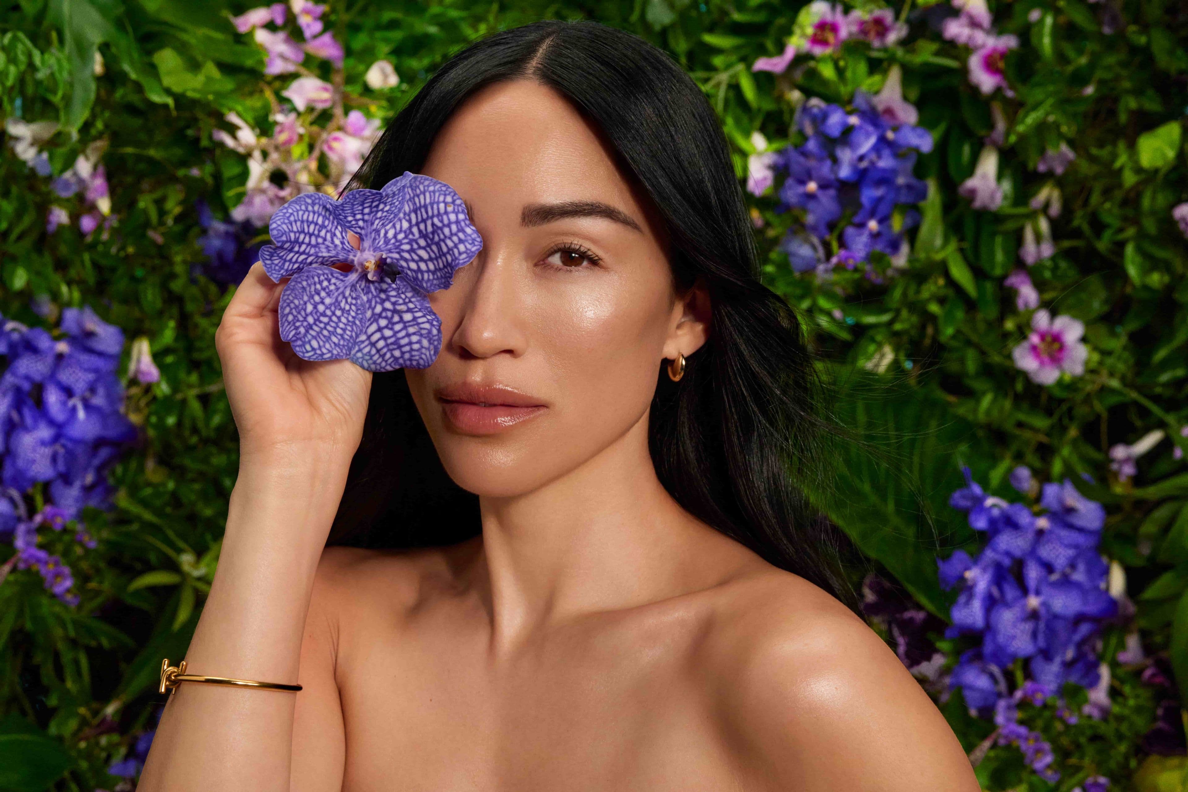 Model holds flowers over right eye with plants and flowers in the background