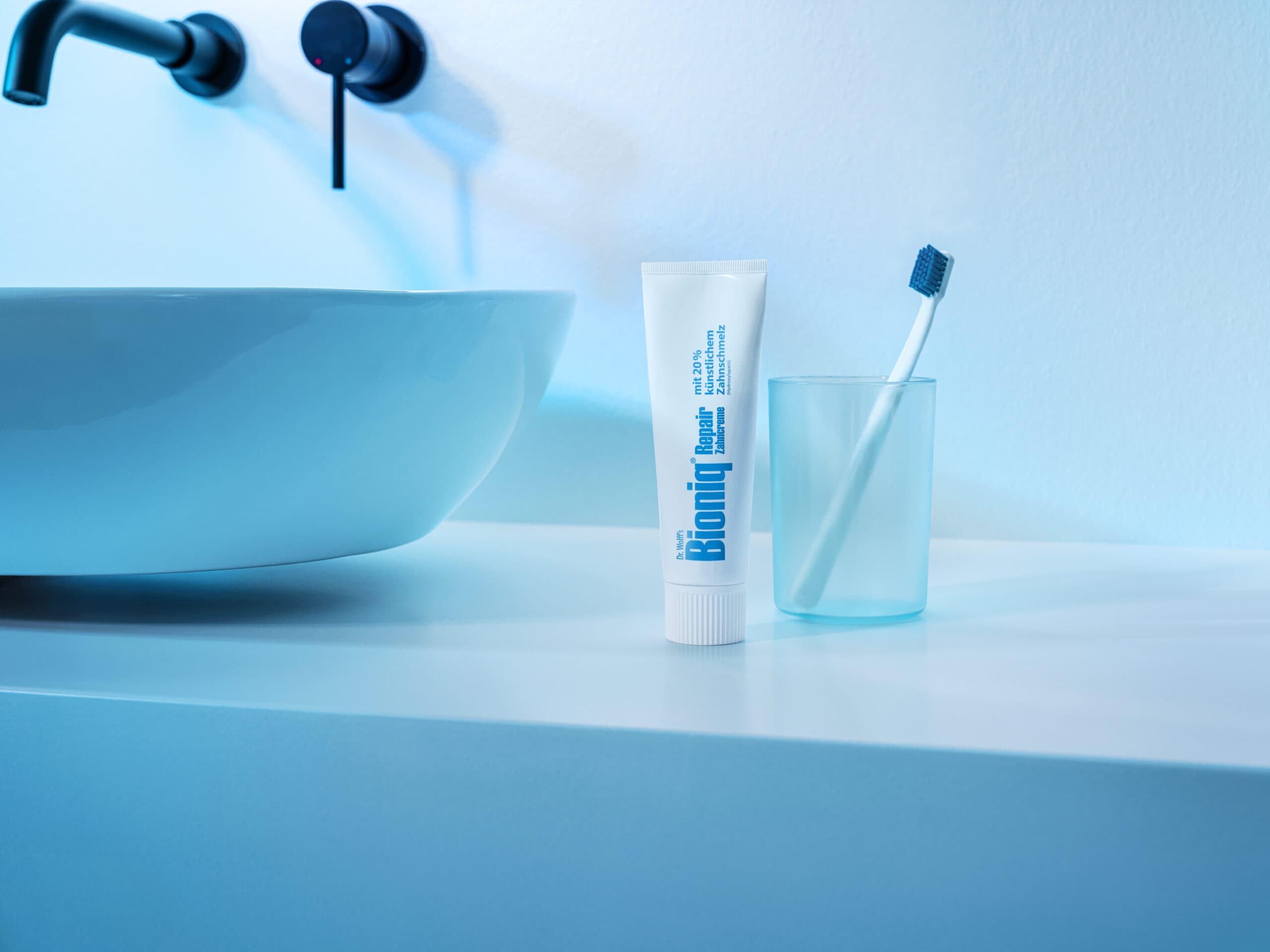 Dr Wolff Bioniq toothpaste tube is displayed on a pale blue surface near a washbasin and glass with white toothbrush.