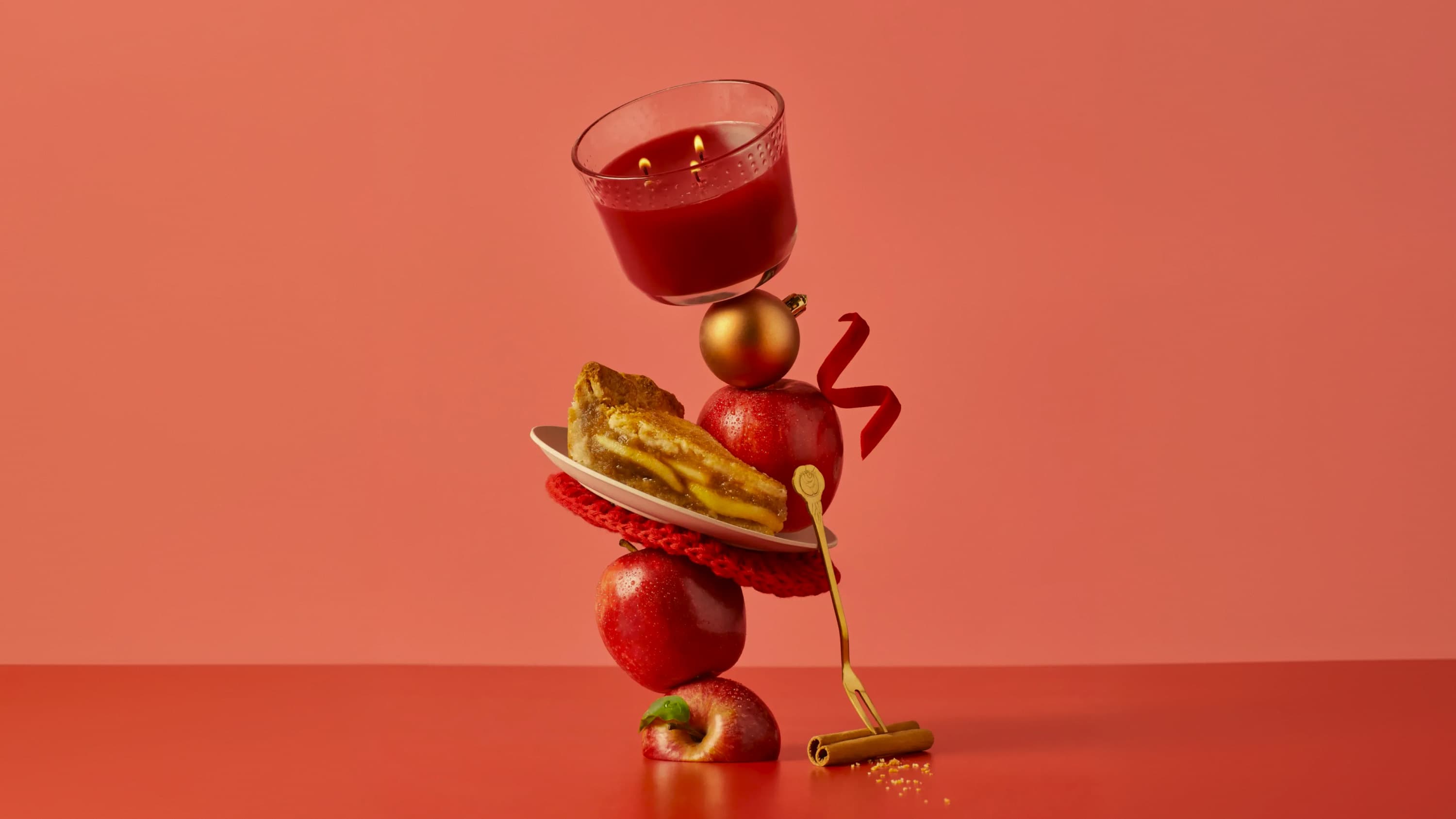A red Glade limited Edition candle is balancing on top of a golden bauble, a piece of apple pie and red apples over a red background.