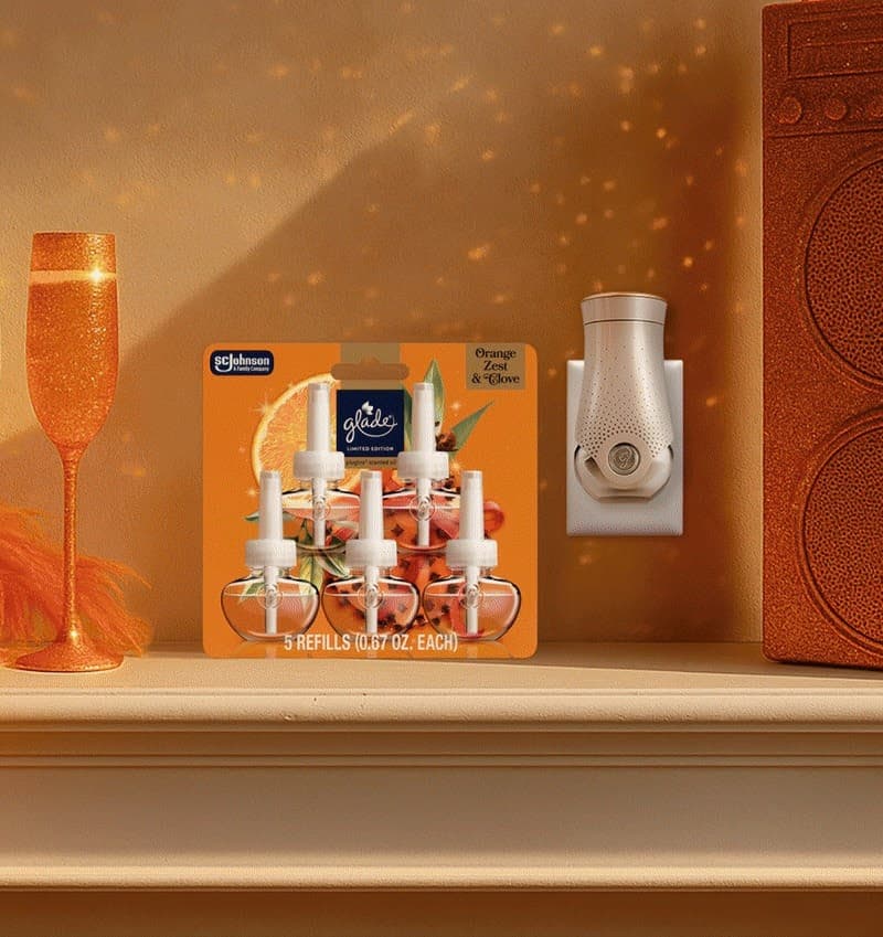 Glade Orange Zest & Clove plug-in air freshener refills on a shelf with a glittery champagne glass and a plugged-in diffuser.